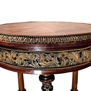 Puede incluir: Una mesa redonda de madera con un acabado marr&oacute;n oscuro y detalles dorados. La mesa tiene un dise&ntilde;o tallado decorativo alrededor del borde y tres patas.