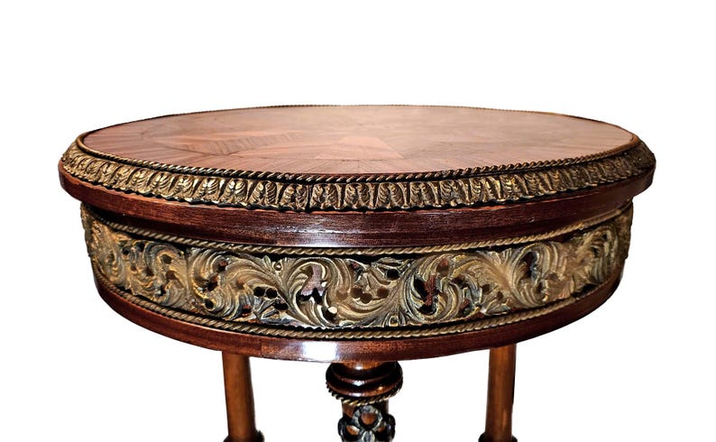 Puede incluir: Una mesa redonda de madera con un acabado marr&oacute;n oscuro y detalles dorados. La mesa tiene un dise&ntilde;o tallado decorativo alrededor del borde y tres patas.