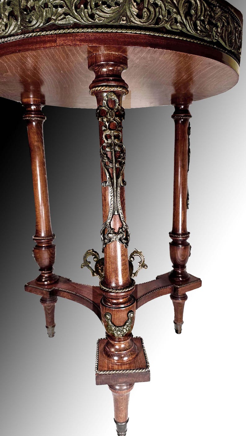 Puede incluir: Una mesa redonda de madera con un borde decorativo de lat&oacute;n alrededor del borde superior. La mesa tiene tres patas con adornos de lat&oacute;n ornamentados y un acabado de madera oscura.