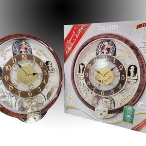 May include: A Seiko Special Collector's Edition musical clock with a brown wooden frame. The clock face features a gold-colored center and silver accents, with decorative figures and Swarovski crystal elements. The clock is displayed next to its packaging.