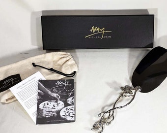 Puede incluir: Un servidor de pastel de plata con un mango negro y un diseño floral. El servidor está empaquetado en una caja negra con una bolsa de polvo blanca y una tarjeta con el texto "Aramy Michael Aram White Orchard Collection".