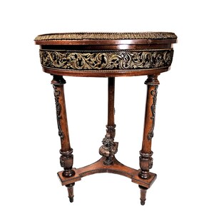 Puede incluir: Una mesa redonda de madera con intrincados tallados alrededor del borde superior. La mesa tiene tres patas con detalles ornamentados y un acabado marr&oacute;n oscuro.