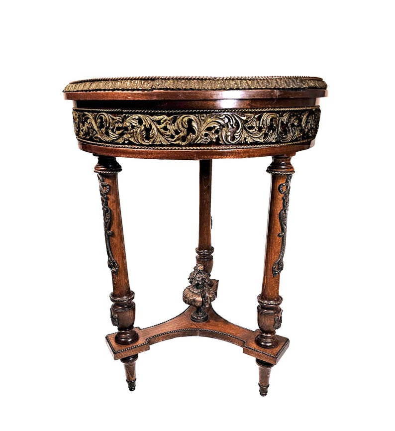 Puede incluir: Una mesa redonda de madera con intrincados tallados alrededor del borde superior. La mesa tiene tres patas con detalles ornamentados y un acabado marr&oacute;n oscuro.