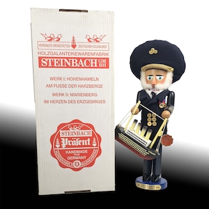 May include: A Steinbach Captain Smith of the Titanic nutcracker figurine, holding a miniature Titanic ship. The figurine is dressed in a navy blue uniform and hat. The box next to it has the Steinbach logo and German text.