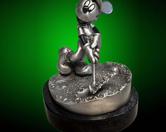Puede incluir: Una figurita de plata de Mickey Mouse vestido con un atuendo de golf, balanceando un palo de golf. La figurita está de pie sobre una base de plata con una base de mármol negro.