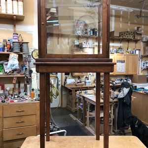 Könnte beinhalten: Ein Vitrinenschrank aus Holz mit Glastüren und einem Holzsockel mit vier Beinen. Die Vitrine ist leer und steht in einer Werkstatt.
