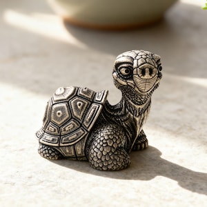 May include: A detailed silver-colored turtle figurine with a textured shell and detailed facial features. The turtle is positioned on a stone surface, with small potted plants in the background. The figurine is a decorative item.