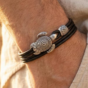 May include: A black leather bracelet with a silver turtle charm and two silver bead accents. The bracelet is made of multiple strands of leather and is secured with a clasp. The turtle charm has intricate detailing.