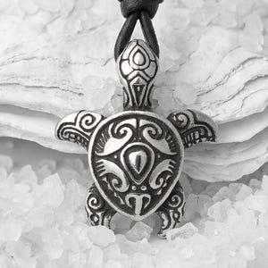 Puede incluir: Collar con colgante de tortuga plateada y cordón de cuero negro. El colgante de tortuga presenta intrincados detalles grabados. El collar se exhibe sobre un lecho de material blanco texturizado.