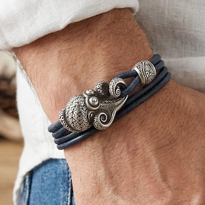 Puede incluir: Pulsera de cuero azul oscuro con cierre de pulpo plateado. La pulsera presenta múltiples hebras de cuero y un diseño detallado de pulpo. La pulsera se lleva en la muñeca, mostrando la intrincada artesanía y el tema náutico.