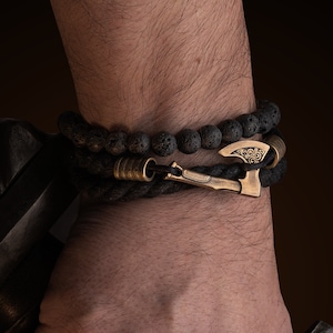 Puede incluir: Dos pulseras en una muñeca. Una está hecha de cuentas de lava negras, la otra es una pulsera de cuerda negra con un cierre dorado en forma de hacha. Las pulseras se usan en una muñeca que sostiene una espada.