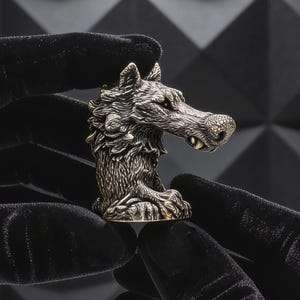 Könnte beinhalten: Eine detaillierte Silberwolfkopf-Figur. Der Wolf ist mit aufwendigem Fell und Gesichtszügen dargestellt. Die Figur wird von behandschuhten Händen vor einem dunklen, strukturierten Hintergrund gehalten. Der Wolfskopf ist etwa 5 cm hoch.