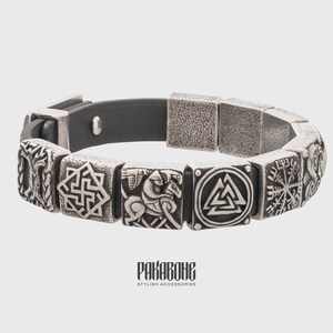 Pode incluir: Um bracelete de couro preto com pingentes de metal prateado que representam v&aacute;rios s&iacute;mbolos n&oacute;rdicos, incluindo um Valknut, um Vegvisir e um cavaleiro a cavalo. O bracelete tem um fecho magn&eacute;tico e o texto "RAKABONE STYLISH ACCESSORIES" est&aacute; impresso na parte inferior.