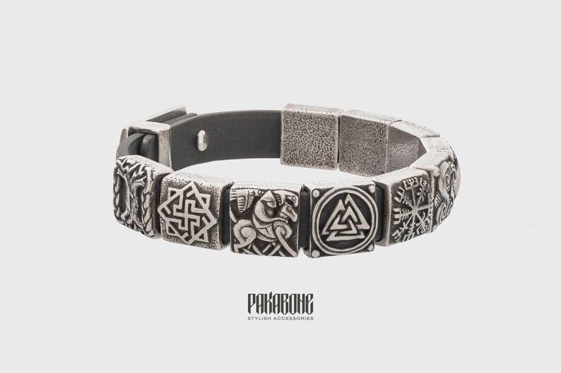 Pode incluir: Um bracelete de couro preto com pingentes de metal prateado que representam v&aacute;rios s&iacute;mbolos n&oacute;rdicos, incluindo um Valknut, um Vegvisir e um cavaleiro a cavalo. O bracelete tem um fecho magn&eacute;tico e o texto "RAKABONE STYLISH ACCESSORIES" est&aacute; impresso na parte inferior.