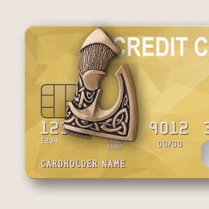 Peut inclure: Une carte de cr&eacute;dit dor&eacute;e avec un charme en m&eacute;tal en forme de hache d&eacute;corative attach&eacute; &agrave; elle. La carte porte l'inscription "CREDIT CARD". Le charme de la hache pr&eacute;sente des motifs complexes de n&oelig;uds.