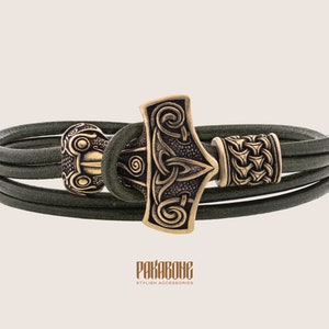 Peut inclure: Un bracelet en cuir vert foncé avec un fermoir de couleur bronze représentant un marteau viking. Le bracelet comporte plusieurs brins de cuir et des accents décoratifs en bronze. La marque "PAKABONE" est visible en bas.