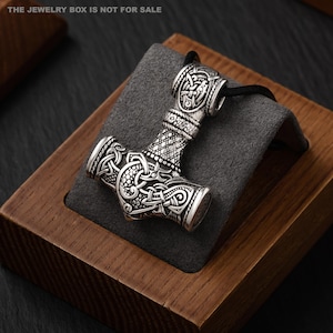 Op de afbeelding: Een zilverkleurige metalen hanger in de vorm van een hamer, voorzien van gedetailleerde reliëfpatronen met knoopwerk en draakachtige motieven. De hanger zit aan een zwart koord en toont bovenaan de tekst THE JEWELRY BOX IS NOT FOR SALE.