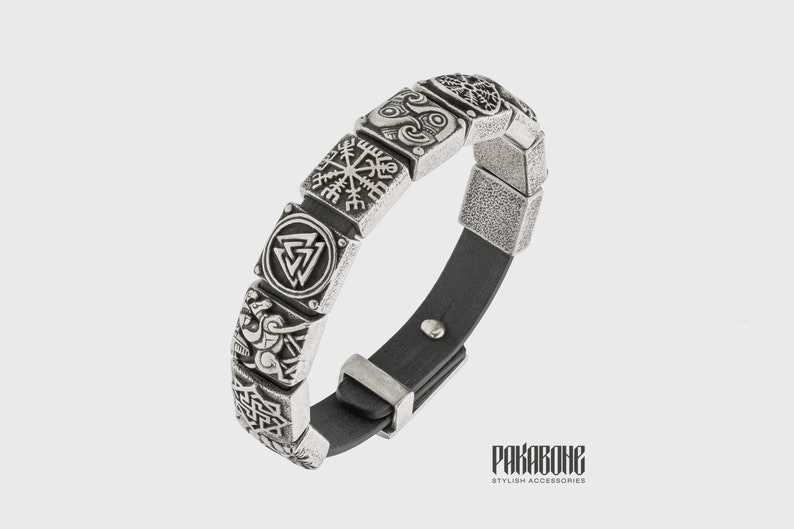 Pode incluir: Uma pulseira de prata com uma tira de couro preto e v&aacute;rios pingentes quadrados com s&iacute;mbolos n&oacute;rdicos. Os pingentes incluem um Vegvisir, um Valknut e um Triskelion. A pulseira tem uma fivela de prata.