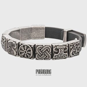 Pode incluir: Uma pulseira de prata com uma tira de couro preto e pingentes de prata com s&iacute;mbolos n&oacute;rdicos. Os pingentes incluem um n&oacute;, um martelo e uma roda.