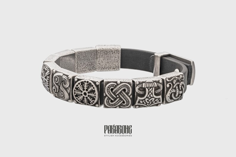 Pode incluir: Uma pulseira de prata com uma tira de couro preto e pingentes de prata com s&iacute;mbolos n&oacute;rdicos. Os pingentes incluem um n&oacute;, um martelo e uma roda.