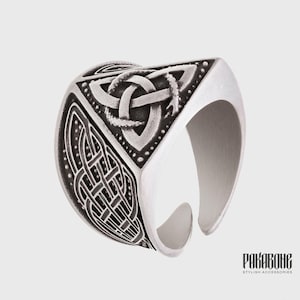 Puede incluir: Un anillo de metal plateado con diseño de banda abierta. La superficie está decorada con un símbolo de nudo triquetra en relieve en la parte superior y complejos patrones de nudos trenzados en los laterales, con detalles oscuros oxidados.