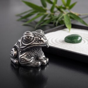 Pode incluir: Uma estatueta de sapo em metal prateado com padrões em relevo intrincados na pele, olhos grandes e uma superfície texturizada. O sapo está posicionado ao lado de uma pequena bandeja preta contendo areia branca e uma pedra verde lisa.