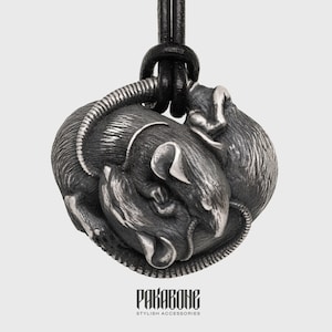 Puede incluir: Un collar con un colgante de plata que presenta tres ratones entrelazados, con un cordón de cuero negro. El colgante está detallado con características realistas y una superficie texturizada.