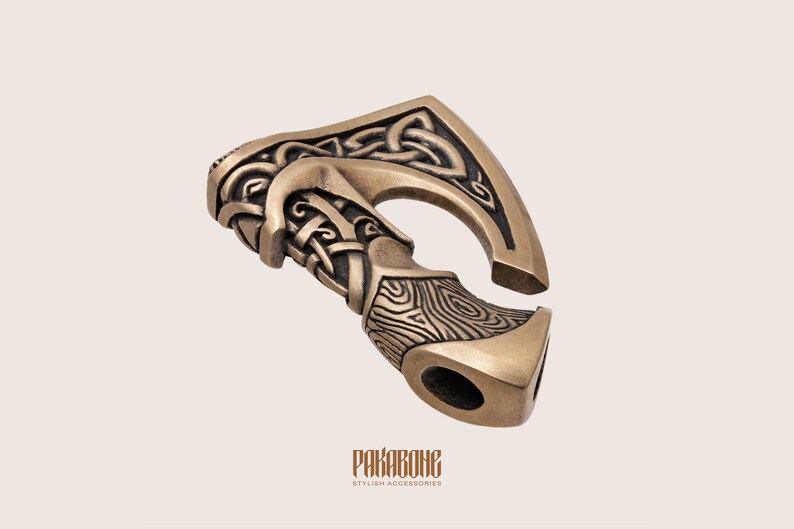 Peut inclure: Un pendentif en bronze en forme de hache avec des d&eacute;tails complexes de n&oelig;uds et de grain de bois. La t&ecirc;te de la hache est incurv&eacute;e et poss&egrave;de un trou pour attacher une cha&icirc;ne ou un cordon. Le pendentif est un accessoire &eacute;l&eacute;gant pour tous ceux qui aiment la culture viking.