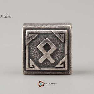 May include: A silver charm with the Othiila rune, a symbol of inheritance and legacy, in a square frame.