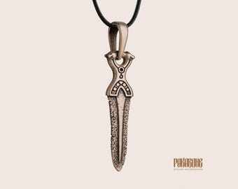 May include: A bronze-toned metal pendant shaped like a dagger, suspended from a black cord. The pendant features an intricate hilt with two animal-like heads at the top and a textured, weathered blade surface. The text RAKABONE is visible at the bottom.