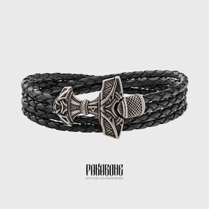 Puede incluir: Pulsera trenzada de cuero negro con un dije de Mjolnir plateado. El dije está decorado con intrincados patrones y tiene una superficie texturizada. La pulsera está hecha de múltiples hebras de cuero que están entrelazadas.