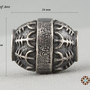 Peut inclure: Une perle argentée avec un design texturé et complexe. La perle mesure 16 mm de long et 13 mm de large, avec une bande centrale et des motifs détaillés sur les extrémités arrondies. Le texte "#6S Helm of Awe" est visible.
