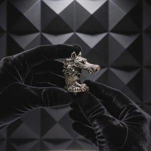 May include: A small, metallic, bronze-toned wolf head figurine featuring detailed fur texture, pointed ears, and a visible tooth. The sculpture is captured in a side profile view, showcasing the intricate craftsmanship of the animal's facial features.