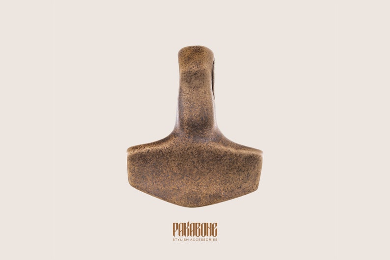 Puede incluir: Un colgante de color bronce con forma de martillo con un bucle en la parte superior. El colgante tiene una superficie texturizada y es un s&iacute;mbolo popular de la mitolog&iacute;a n&oacute;rdica.