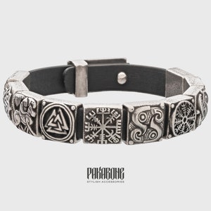 Pode incluir: Um bracelete de couro preto com pingentes de metal prateado que representam v&aacute;rios s&iacute;mbolos n&oacute;rdicos, incluindo o Valknut, o Vegvisir e o s&iacute;mbolo do deus Odin. O bracelete tem um fecho de fivela.