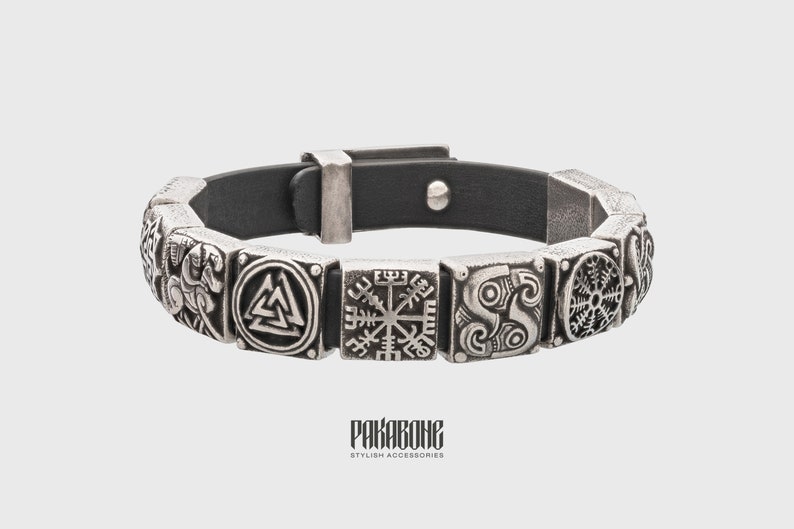 Pode incluir: Um bracelete de couro preto com pingentes de metal prateado que representam v&aacute;rios s&iacute;mbolos n&oacute;rdicos, incluindo o Valknut, o Vegvisir e o s&iacute;mbolo do deus Odin. O bracelete tem um fecho de fivela.