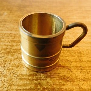 Könnte beinhalten: Eine kleine, goldfarbene Metalltasse mit Henkel. Die Tasse ist fassförmig und hat eine glatte, glänzende Oberfläche.
