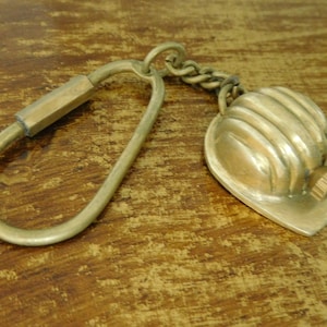 Könnte beinhalten: Ein Schlüsselanhänger aus Messing mit einem Miniatur-Schutzhelm. Der Schlüsselanhänger hat einen einfachen ovalen Ring und eine kleine Kette, die den Helm verbindet.
