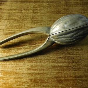 Könnte beinhalten: Ein silberfarbener Nussknacker in Form einer Walnuss mit einer Scharnieröffnung. Der Nussknacker hat zwei lange Arme, die sich aus der Walnussschale erstrecken.