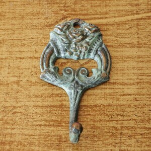 Puede incluir: Un gancho de metal decorativo con un diseño floral. El gancho es de color verde claro con una pátina marrón. Está montado en una superficie de madera.