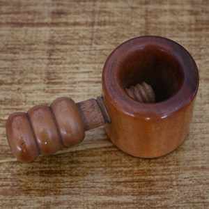 Könnte beinhalten: Ein Nussknacker aus Holz mit brauner Oberfläche. Der Nussknacker hat eine Gewindeschraube in der Mitte und einen Griff mit einem abgerundeten, strukturierten Griff.