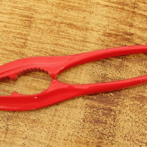 Könnte beinhalten: Ein roter Nussknacker aus Metall mit einem strukturierten Griff und einer gezahnten Kante. Der Nussknacker ist zum Knacken von Nüssen gedacht und ein Küchenutensil.