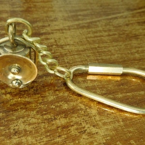 Può includere: Portachiavi in ottone in stile vintage con un piccolo ciondolo dettagliato a forma di sistema di pulegge. Il ciondolo ha un disco circolare color rame ed è collegato a una catena e a un portachiavi grande a forma ovale.