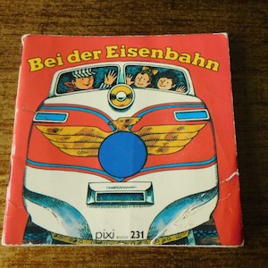 Peut inclure: Un livre pour enfants vintage intitulé "Bei der Eisenbahn" avec une couverture rouge. La couverture représente un train de dessin animé avec des passagers qui saluent depuis les fenêtres. Le livre porte le numéro 231 et le mot "pixi" est imprimé en bas.