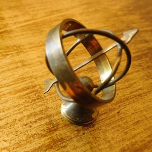 Könnte beinhalten: Ein Messing-Desktop-Ornament in Form einer sich drehenden Kugel mit einem kleinen Pfeil, der oben befestigt ist. Die Kugel steht auf einem runden Sockel.