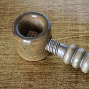 Könnte beinhalten: Ein Nussknacker aus Holz mit brauner Oberfläche. Der Nussknacker hat einen runden, ausgehöhlten Bereich zum Knacken von Nüssen und einen Griff mit einer Gewindeschraube.