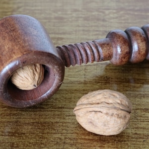 May include: A wooden nutcracker with a brown finish and a single walnut inside the cracking mechanism. A whole walnut is also visible on the table.