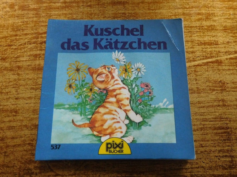 Puede incluir: Un libro azul con el t&iacute;tulo "Kuschel das K&auml;tzchen" y una imagen de un gatito naranja y blanco mirando hacia arriba a una flor. El libro forma parte de la serie "Pixi B&uuml;cher" y tiene el n&uacute;mero 537 en la portada.