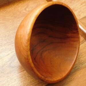 May include: A large, wooden bowl with a smooth, rounded interior. The bowl is made of light brown wood with a rich grain pattern.