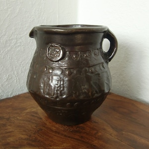 May include: A dark brown ceramic jug with a handle and a raised design. The jug has a circular stamp on the front.